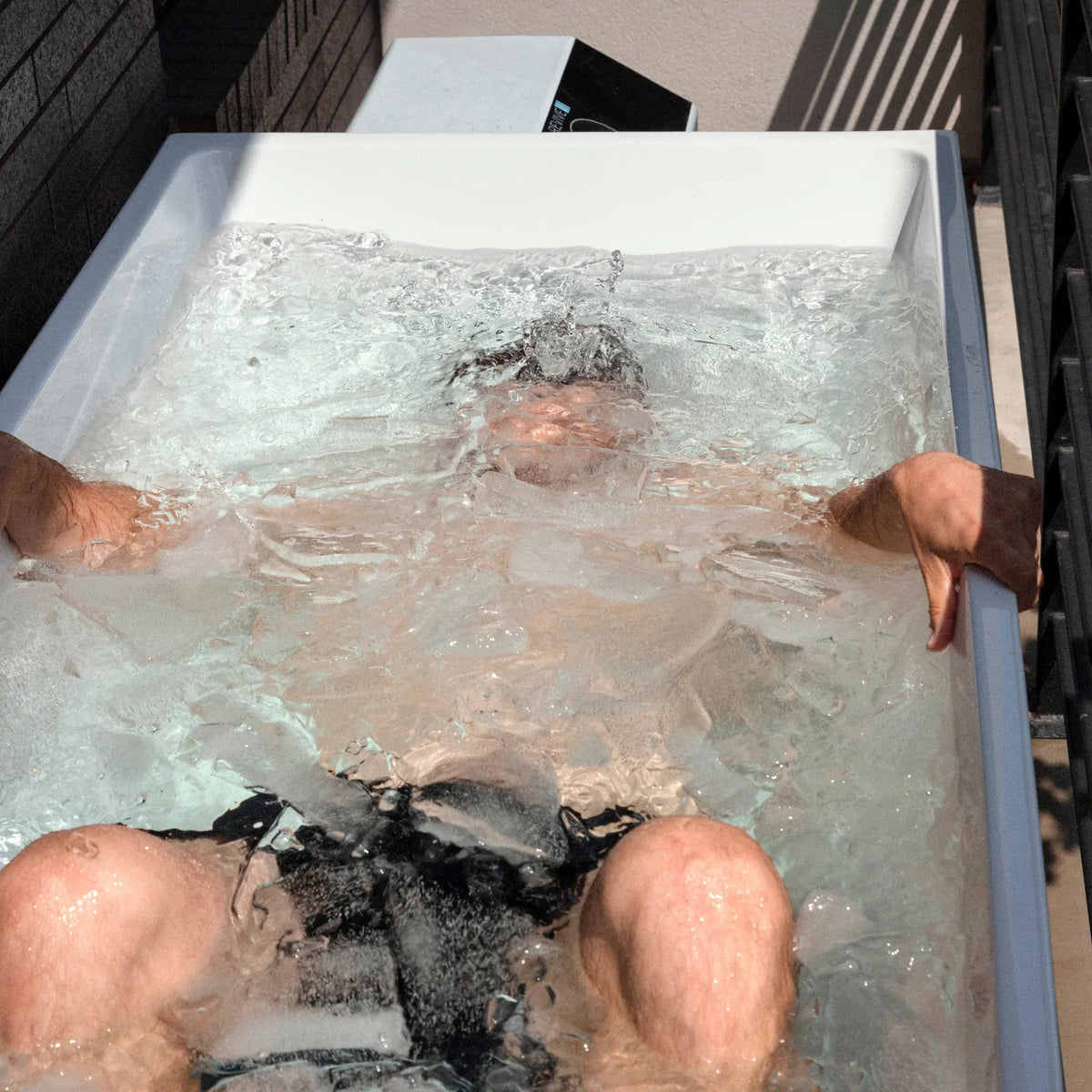 Person reclining in a white Revive acrylic cold plunge with water and floating ice visible